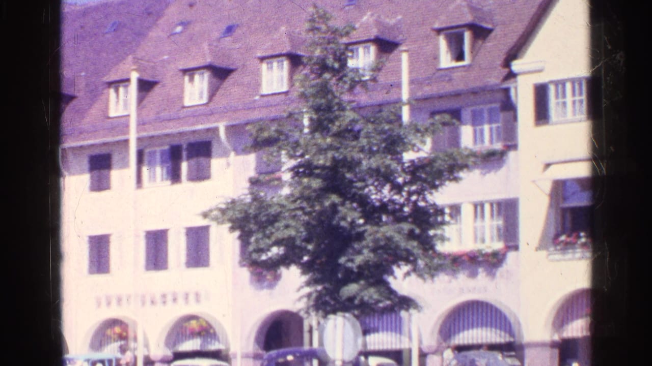 1961 Switzerland street frame showing shuttered facades, a leafy tree, striped awnings, flower boxes, and arched lower-level openings.