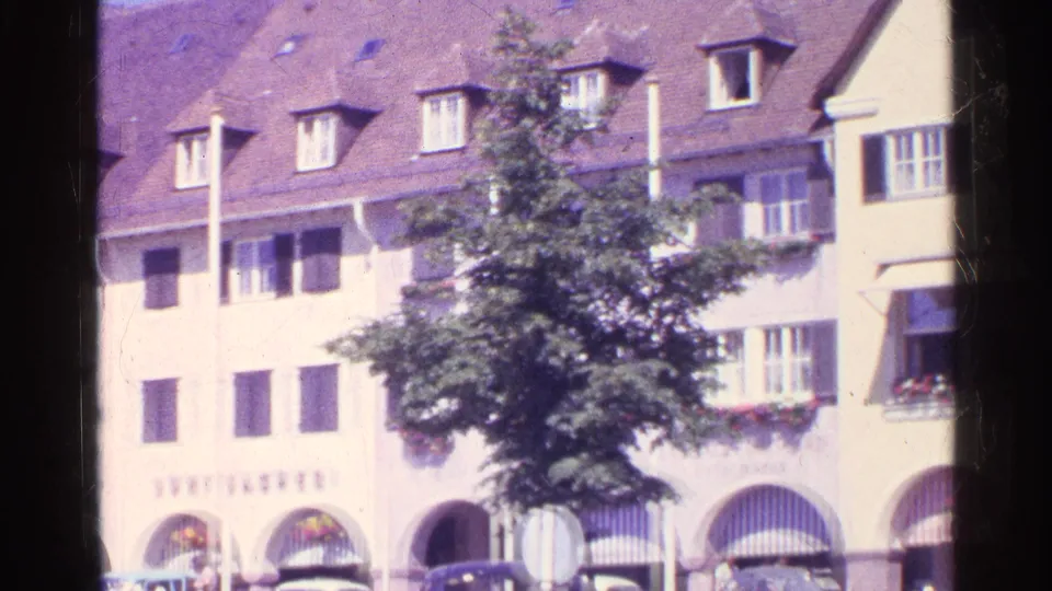 1961 Switzerland street frame showing shuttered facades, a leafy tree, striped awnings, flower boxes, and arched lower-level openings.