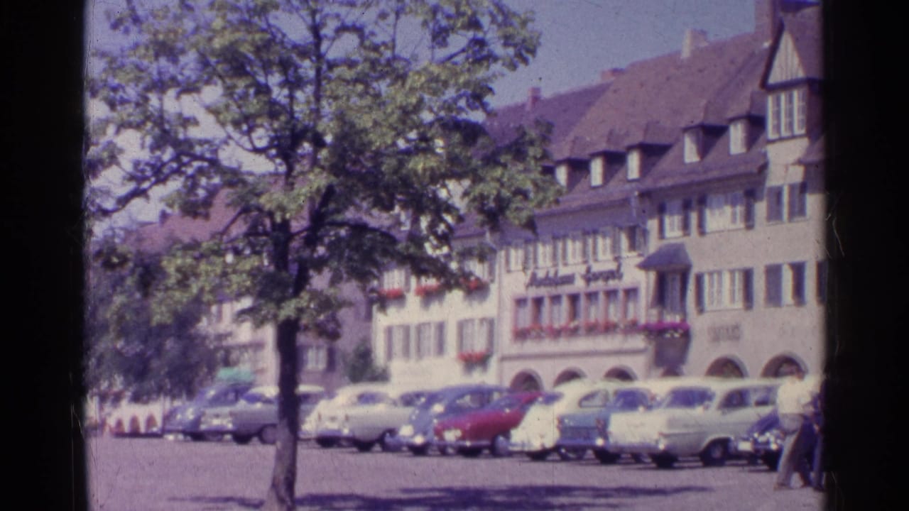 1961 Swiss town square view with parked mid-century cars, a tree, arched storefronts, and gabled buildings seen from a handheld 8mm camera.