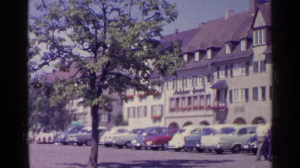 1961 Swiss town square view with parked mid-century cars, a tree, arched storefronts, and gabled buildings seen from a handheld 8mm camera.
