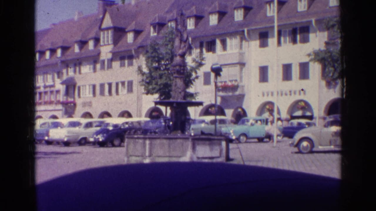 1961 Swiss square with a central stone fountain, parked classic cars, arched shopfronts, and traditional multi-story buildings.
