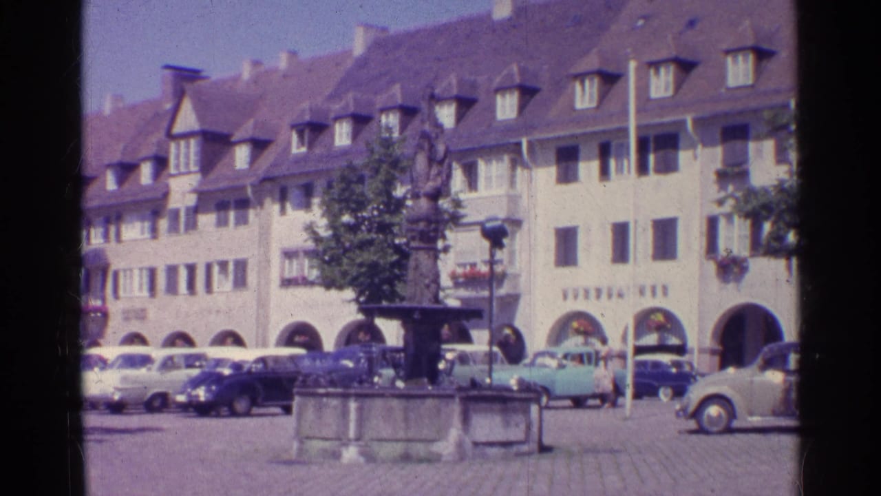 1961 Swiss town-square frame showing a statue fountain, classic cars, arched storefronts, gabled roofs, and a pedestrian at the edge.