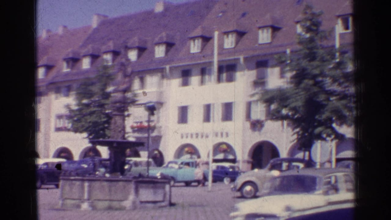 Final 1961 Swiss town-square view with a stone fountain, parked classic cars, trees, arched storefronts, and softly blurred 8mm motion.