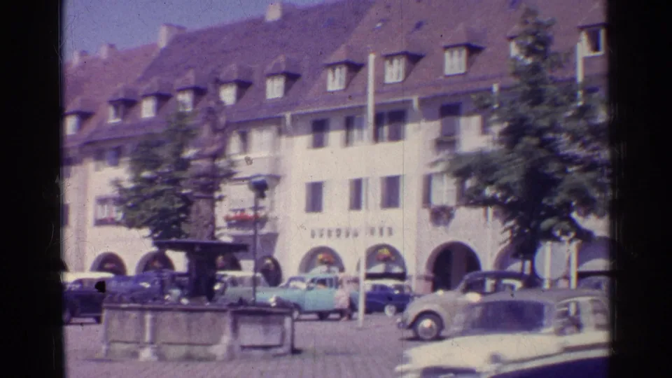 Final 1961 Swiss town-square view with a stone fountain, parked classic cars, trees, arched storefronts, and softly blurred 8mm motion.
