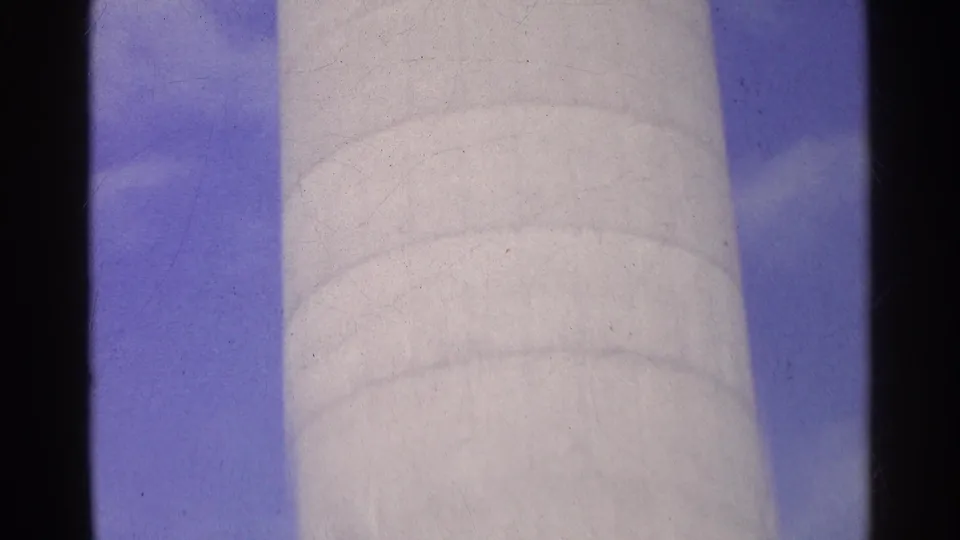 1961 Switzerland 8mm frame showing a pale cylindrical tower or column viewed upward against blue sky and clouds.