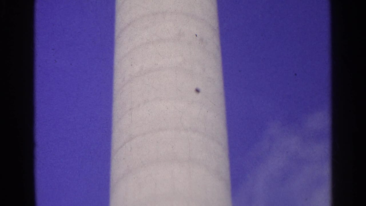 Swiss 1961 home-movie frame with a light cylindrical vertical structure, blue sky, clouds, and visible film texture.