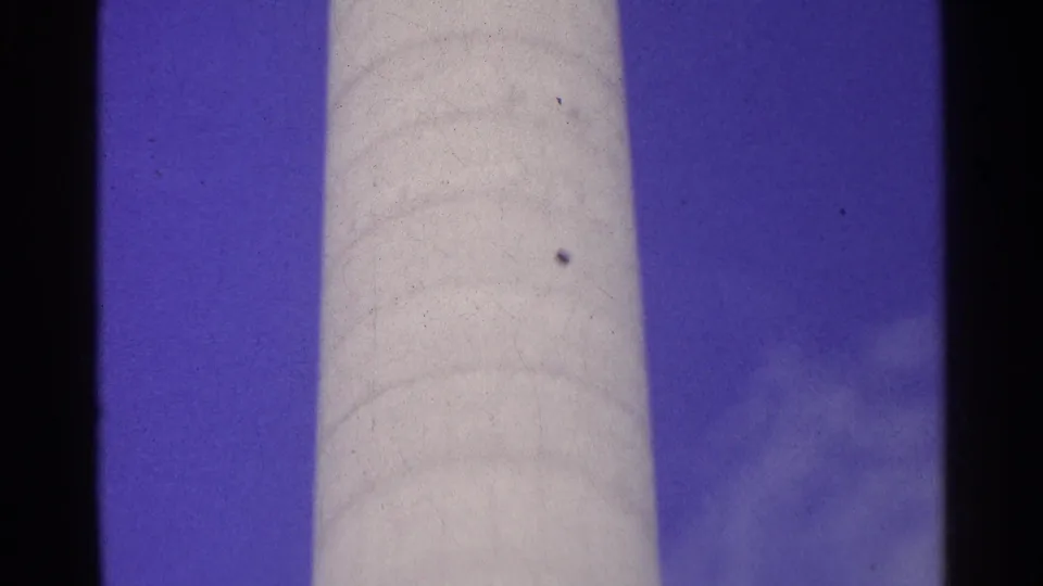 Swiss 1961 home-movie frame with a light cylindrical vertical structure, blue sky, clouds, and visible film texture.