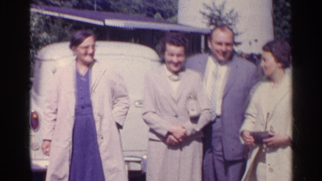 Well-dressed adults smile and gather beside a pale car in this 1961 Switzerland home-movie frame.