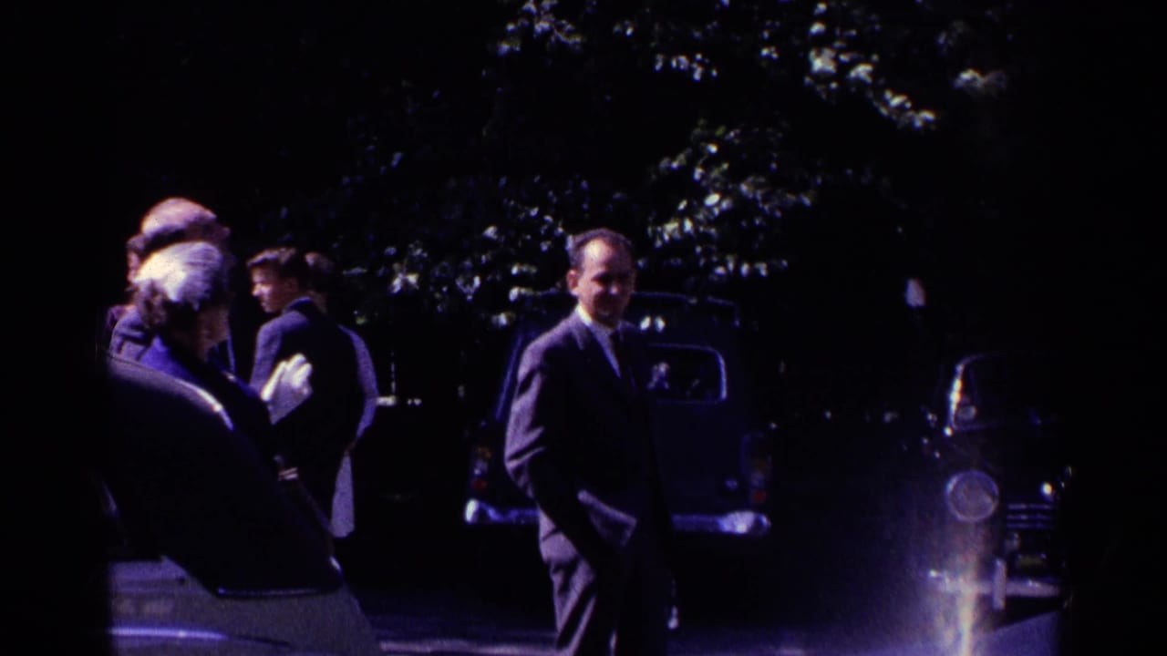 Shaded 1961 Switzerland home-movie frame with adults standing near parked cars beneath trees.