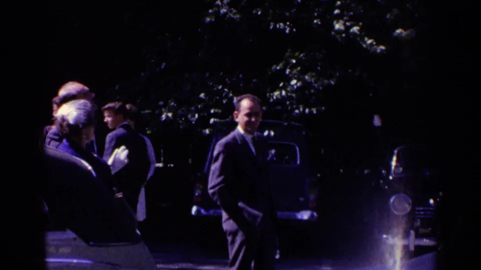 Shaded 1961 Switzerland home-movie frame with adults standing near parked cars beneath trees.