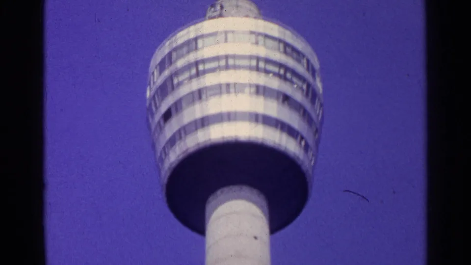 1961 Switzerland home-movie frame showing the white shaft and circular deck of a tall observation tower against a saturated blue sky.