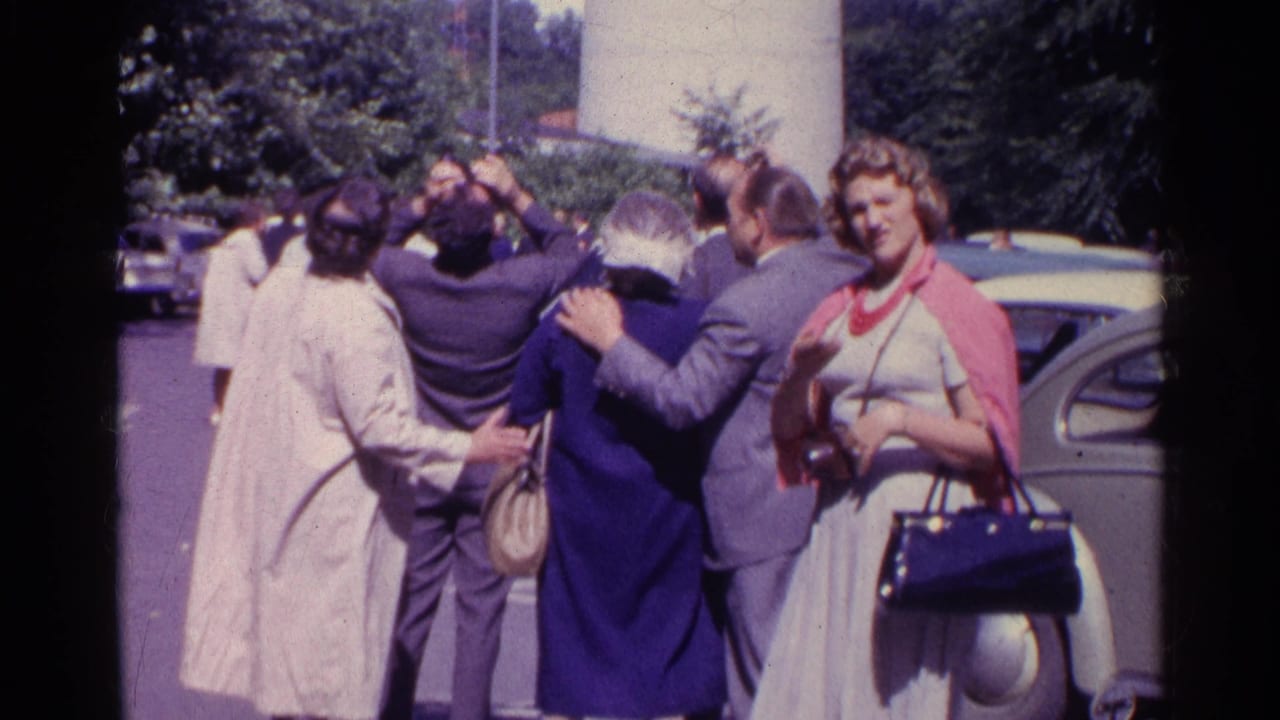 Men and women in 1961 Switzerland gather below the observation tower near cars and trees, with visible faces and some people looking up.