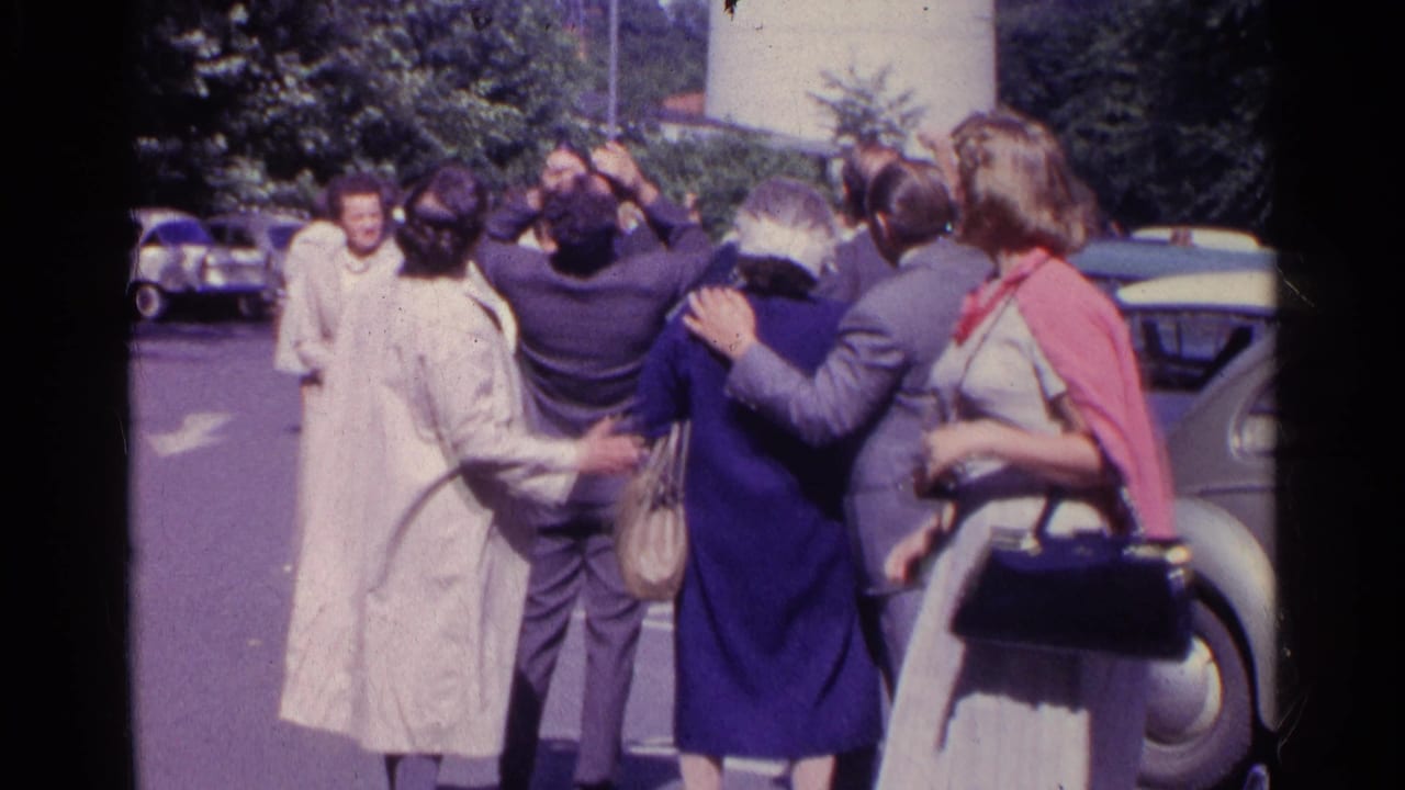 Adults in mid-century clothing gather beside parked cars and look upward.