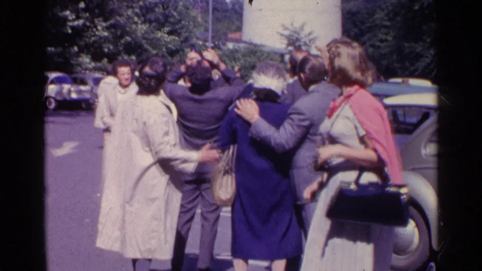 In 1961 Switzerland, adults in coats, suits, and dresses stand near vintage cars while looking up together.