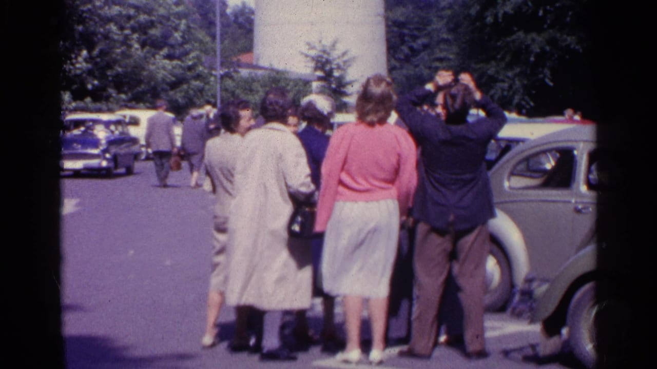 Adults in 1960s attire stand in a Swiss parking area near a compact car, with some looking upward.