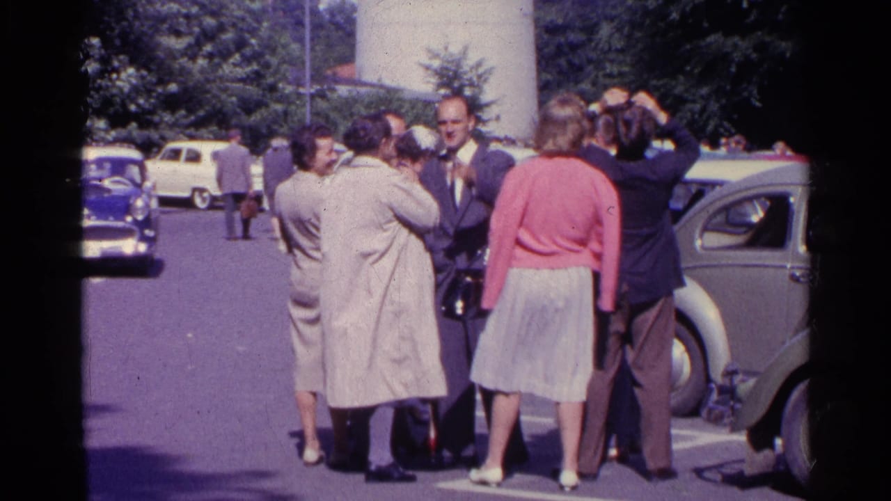 A Swiss 1961 home-movie frame shows adults in coats and suits near parked vintage cars, some still reacting upward.