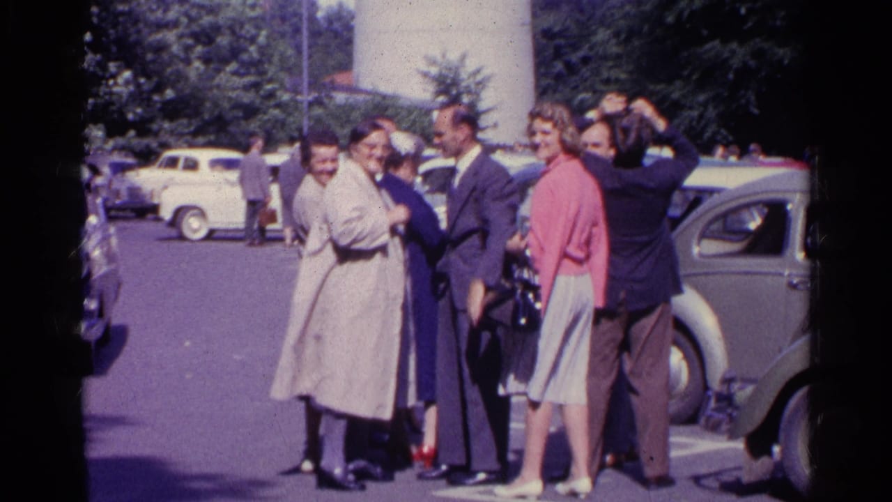 Adults in 1961 Switzerland stand together near a Beetle-shaped car, with visible faces and skyward attention.
