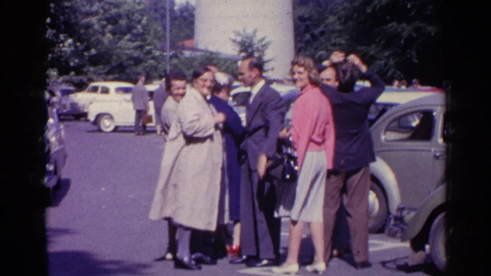 Adults in 1961 Switzerland stand together near a Beetle-shaped car, with visible faces and skyward attention.