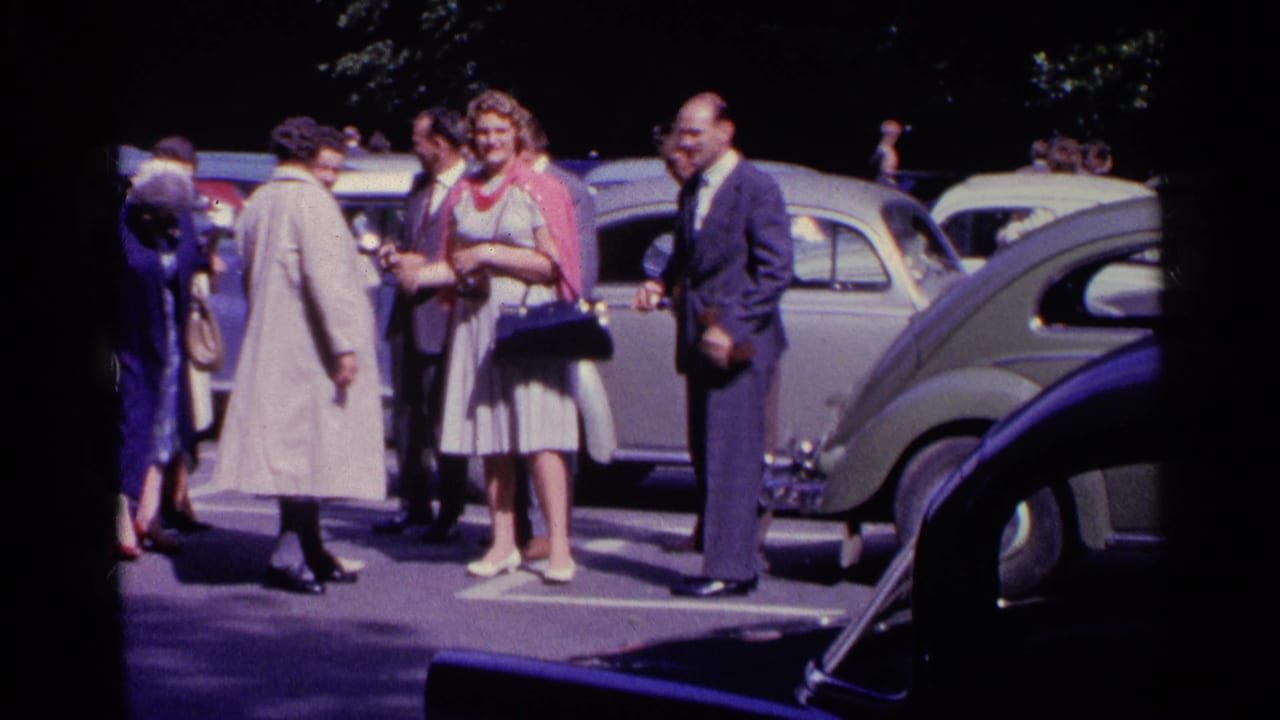 In 1961 Switzerland, well-dressed adults gather and talk beside classic cars in a sunlit parking lot.