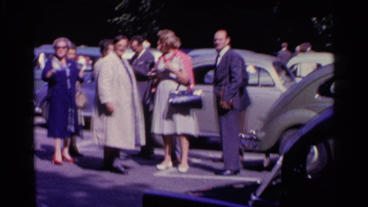 Well-dressed adults in 1961 Switzerland stand in conversation among parked cars, including a light-colored Beetle-like car.