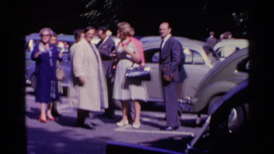 Well-dressed adults in 1961 Switzerland stand in conversation among parked cars, including a light-colored Beetle-like car.