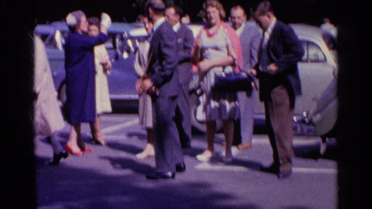 Adults in formal clothing move through a Swiss parking lot while one woman adjusts her hat or hair near parked cars.