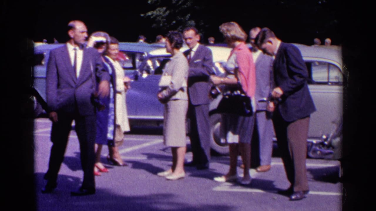 A suited man faces the camera while adults converse near a light-colored Volkswagen Beetle in 1961 Switzerland.