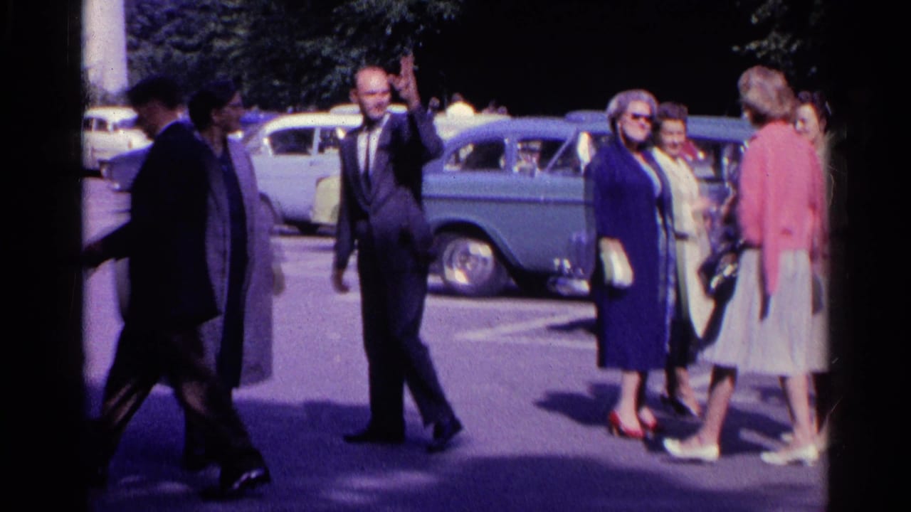 1961 Switzerland frame of adults moving through a parking area with rounded mid-century cars in the background.