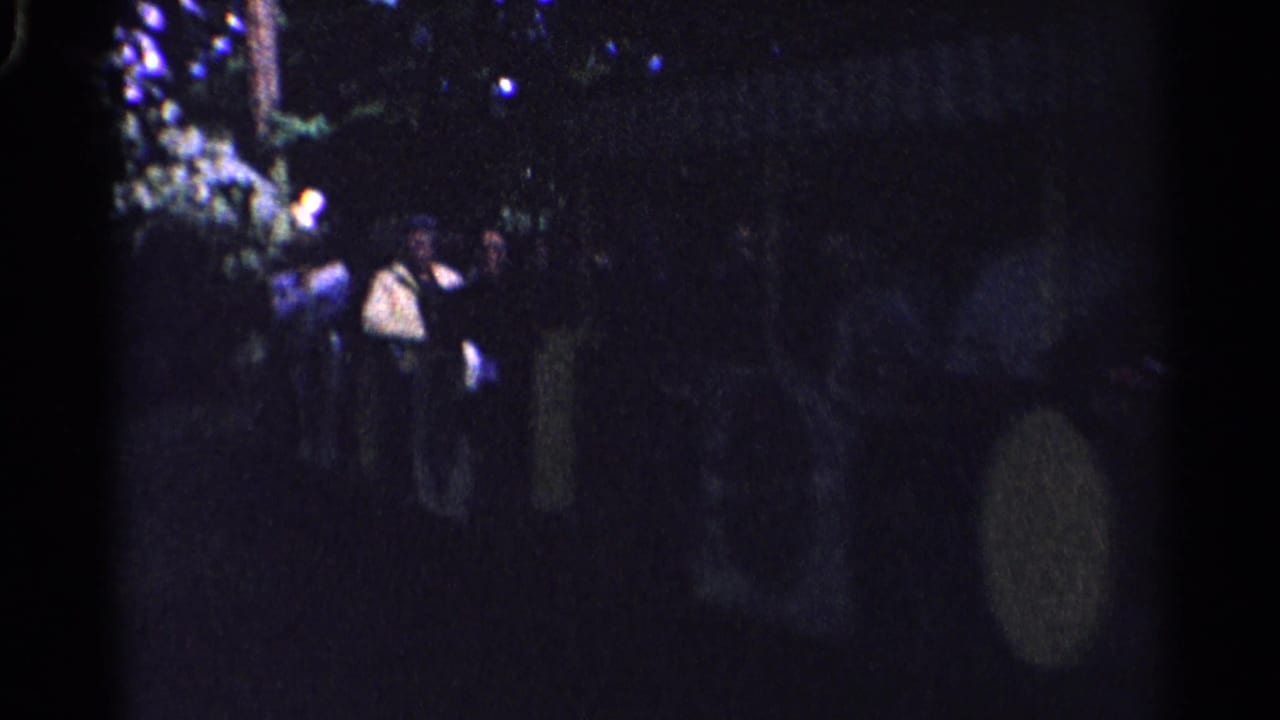 1961 Switzerland home-movie frame with several people standing in a dark outdoor setting near foliage and lights.