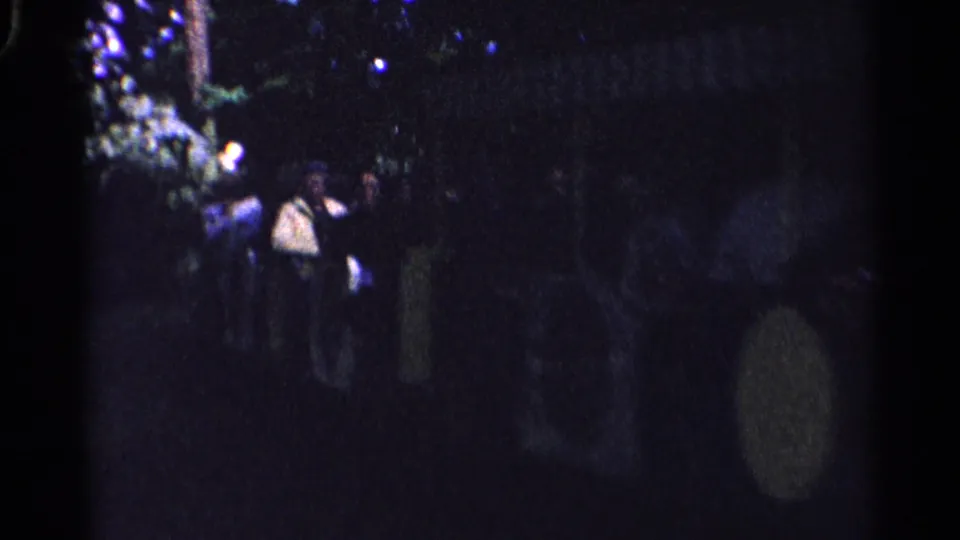 1961 Switzerland home-movie frame with several people standing in a dark outdoor setting near foliage and lights.
