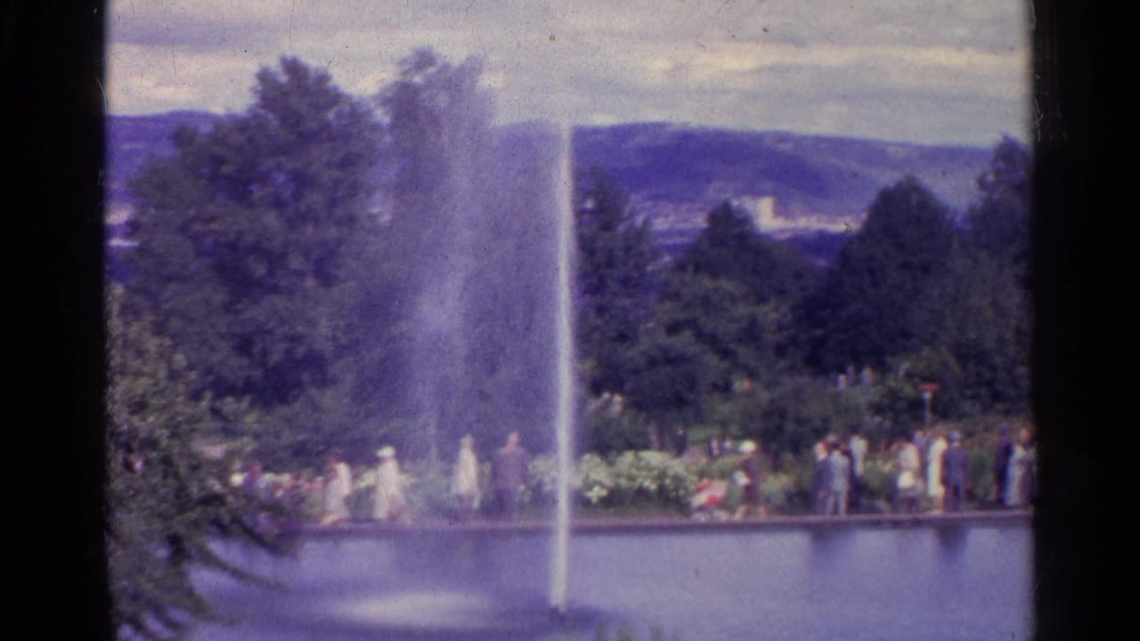 Park pond with a tall fountain, visitors on a path, trees, and distant hills.