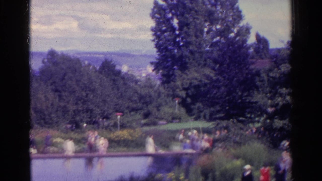 1961 Swiss garden view with visitors near a pond, flowerbeds, dense trees, and a distant hillside.