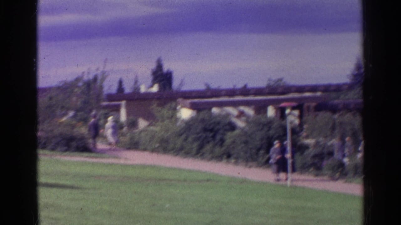 People walking on a garden path beside lawns and shrubs under an overcast sky.