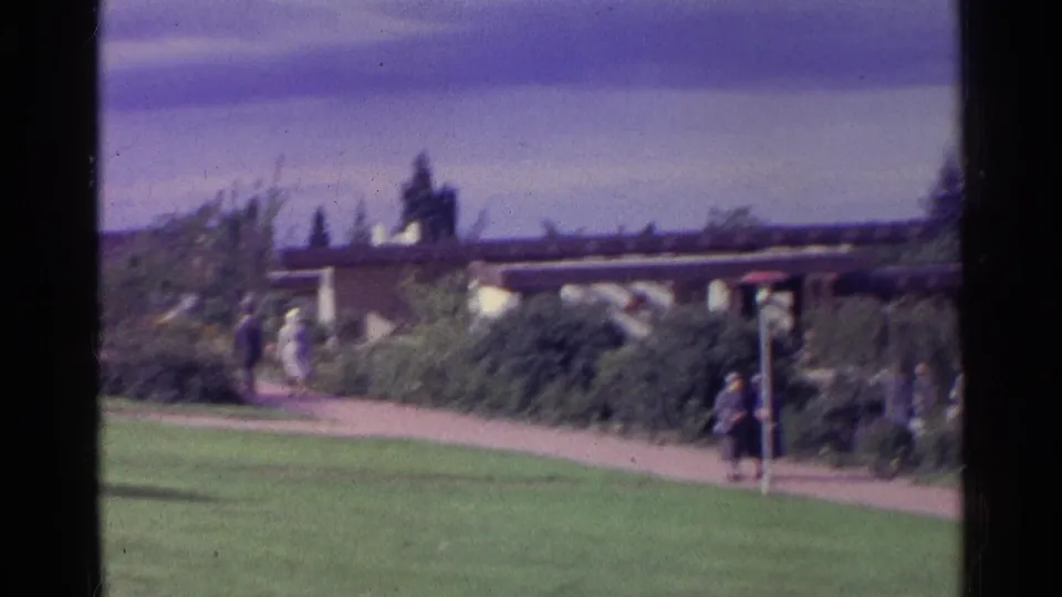 1961 Swiss 8mm frame showing distant people on a curving garden path near lawns, shrubs and a flat-roof structure.