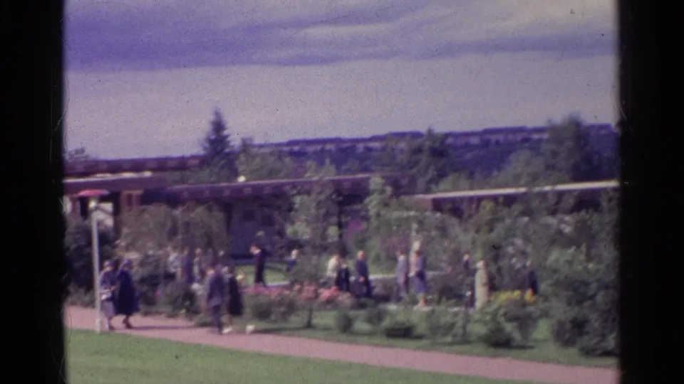 1961 Swiss home-movie frame with groups of visitors on a path bordered by flowers, shrubs and lawn near a low roofline.