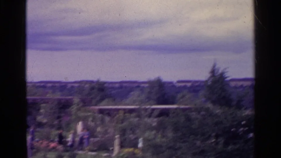 Soft 1961 Swiss 8mm frame showing a landscaped garden with shrubs, distant people, a path and a low modernist roofline.
