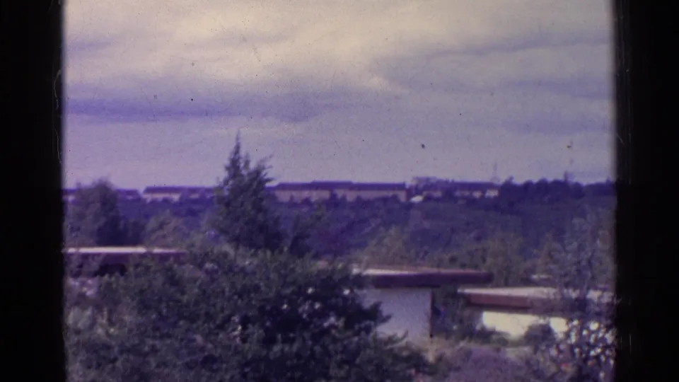 1961 Switzerland home-movie frame with trees, flat-roofed structures, distant ridge, and the dark vertical edge of the film frame.