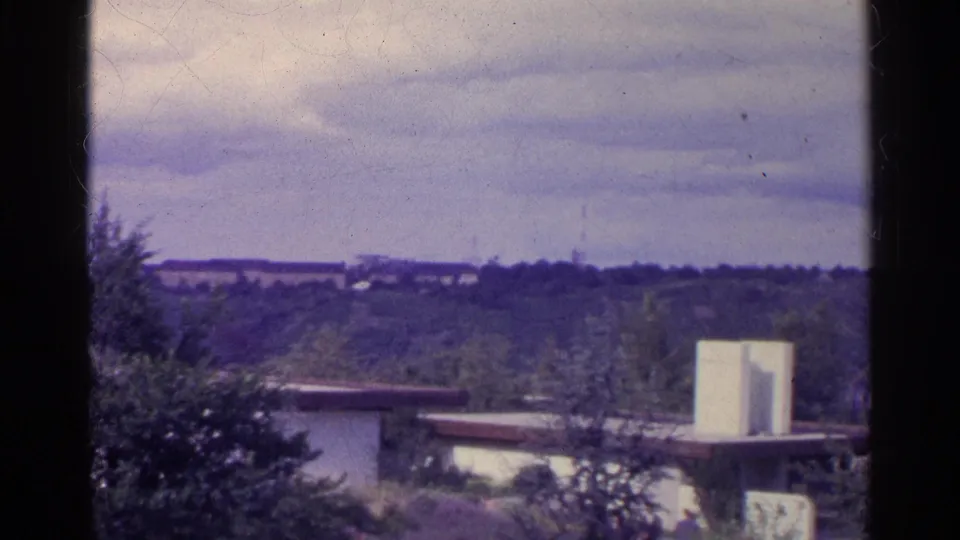 1961 Switzerland frame showing a flat-roofed building, white tower-like form, dense plantings, and wooded hills in the distance.