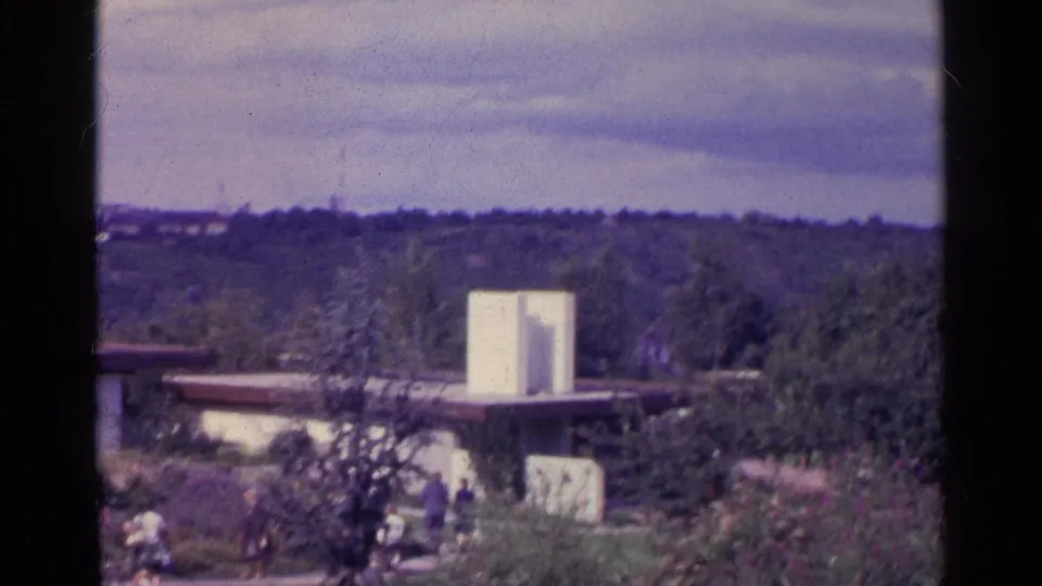 1961 Switzerland 8mm frame of a gardened compound with a flat-roofed building, white structure, trees, and small people outdoors.