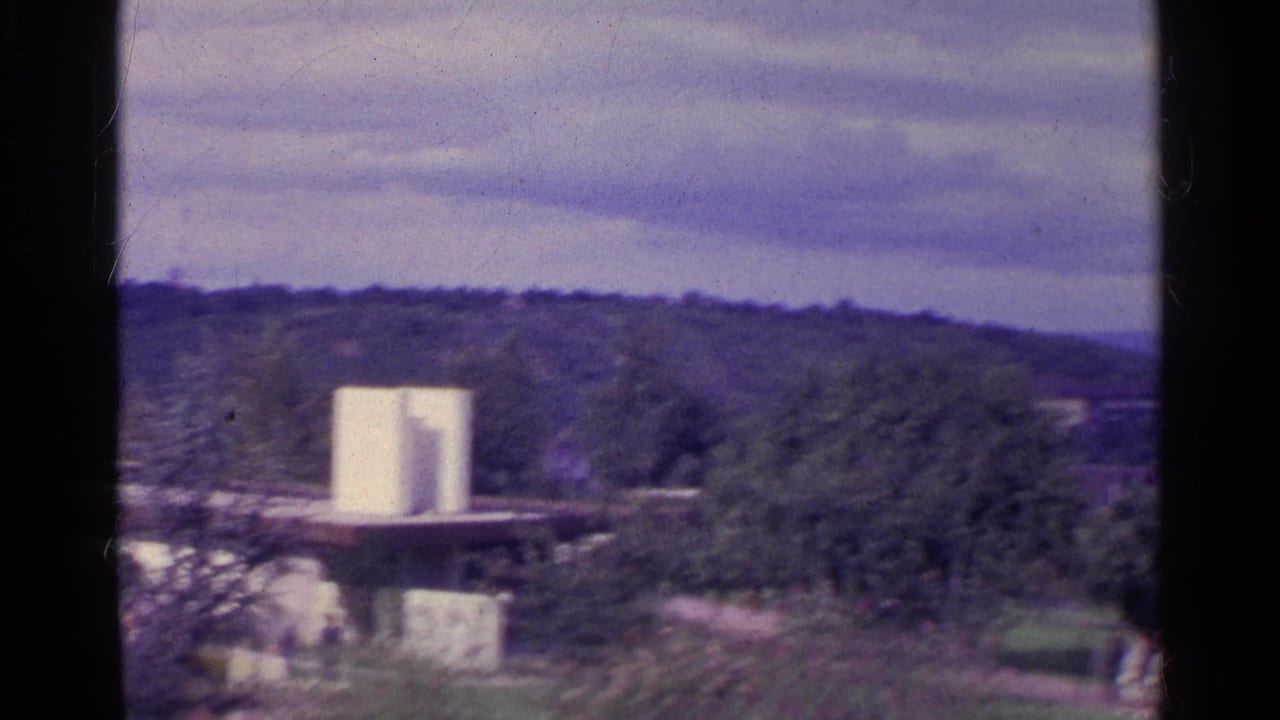 1961 Swiss 8mm frame showing trees, low hills, a pale modern structure, and distant people walking near greenery.