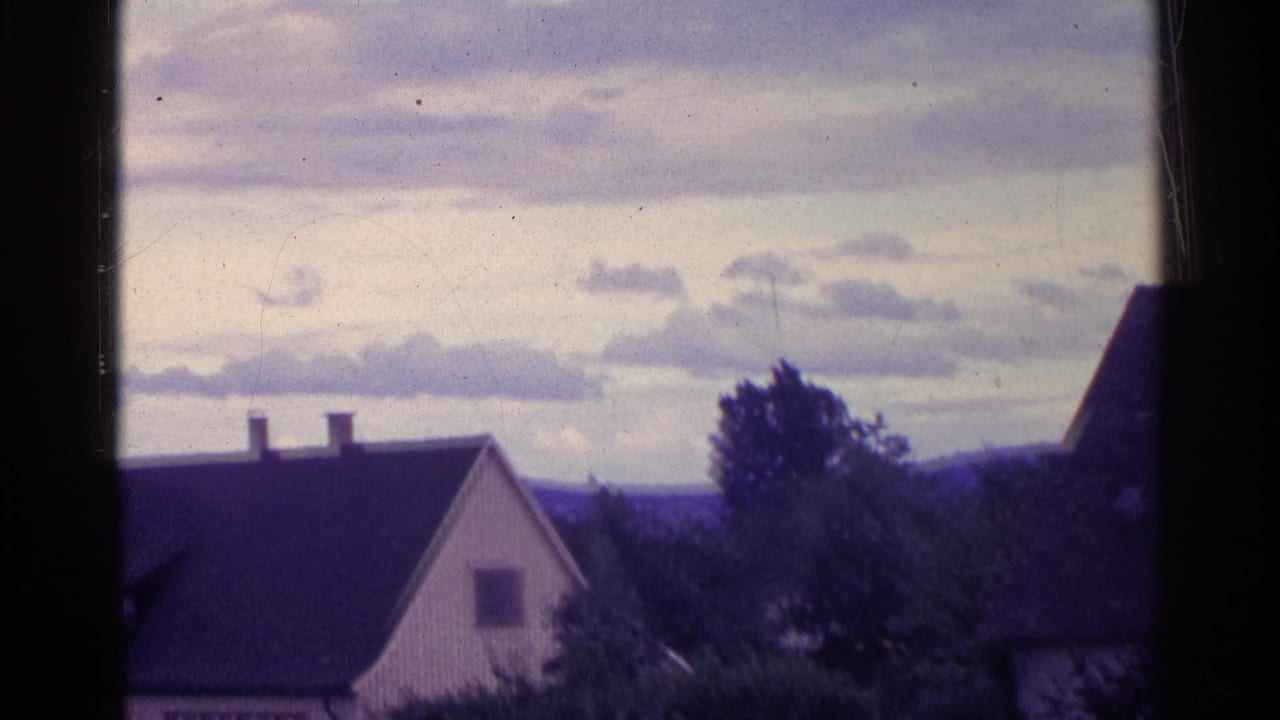 1961 Swiss 8mm frame with a steep-roofed house, two chimneys, trees, distant hills, and a cloudy sky.