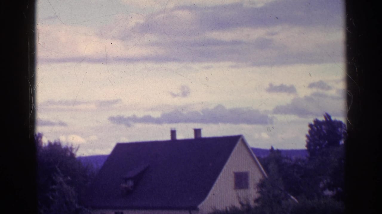 1961 Swiss home-movie frame showing a dark house roof, two chimneys, nearby trees, clouds, and distant hills.