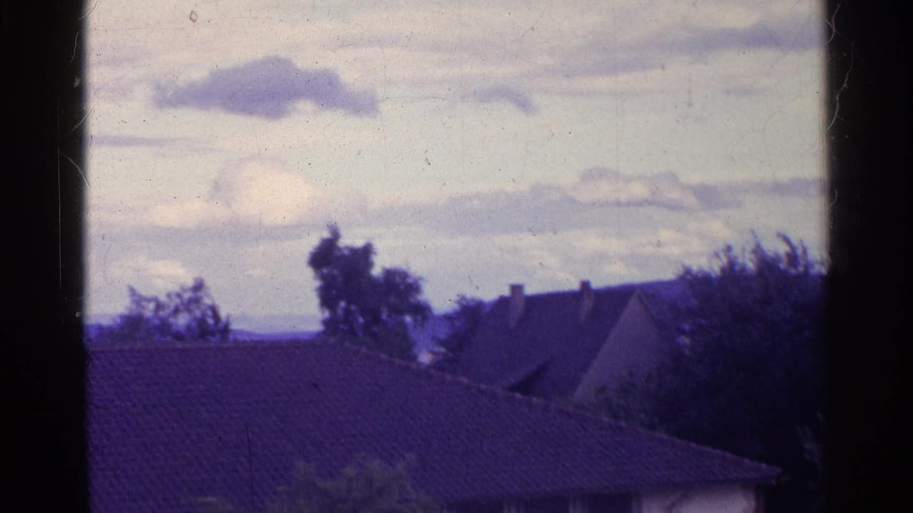 View past a dark window frame toward rooftops, trees, hills, and cloudy sky.