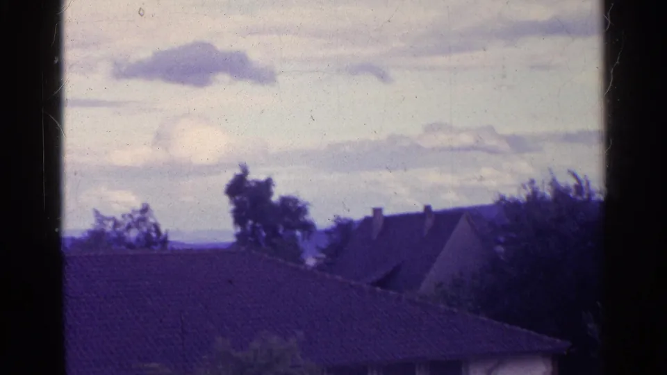 1961 Swiss 8mm view from a window toward tiled rooftops, trees, distant hills, and a cloudy sky.