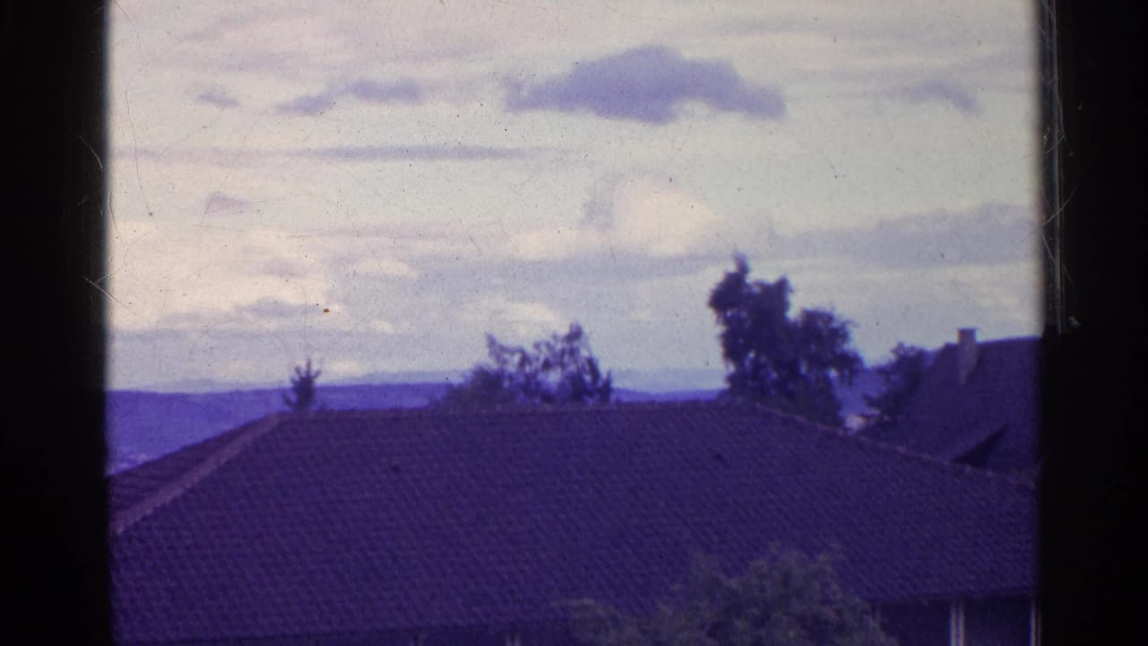 Rooftop in foreground with trees, distant hillside, and cloudy sky beyond.