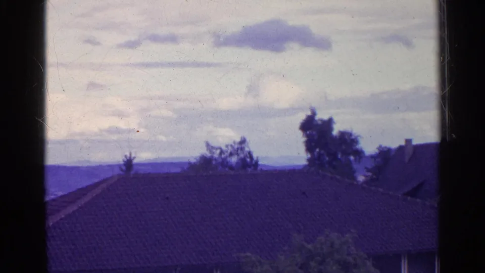 Swiss home-movie frame showing a foreground tiled roof, trees, distant hills, and pale cloud cover.