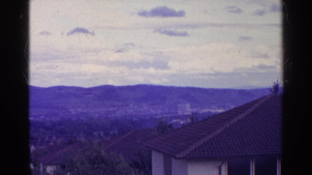 Hazy town rooftops below purple-blue mountains under a cloudy sky.