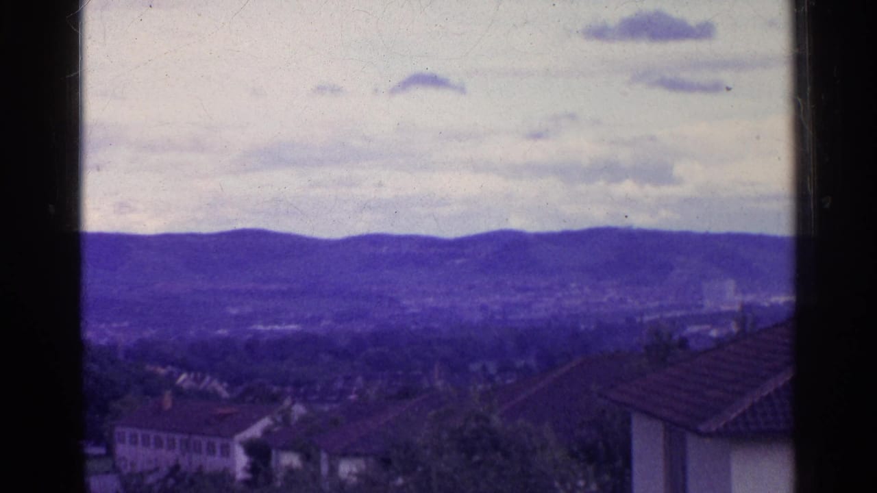 A Swiss town appears below distant purple hills, with a tiled roof in the foreground and dark 8mm frame borders visible.