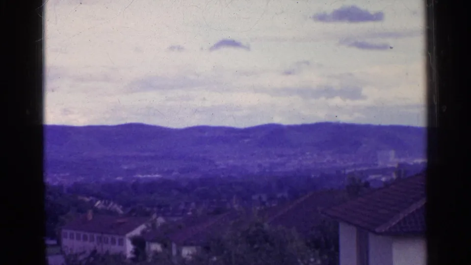 A Swiss town appears below distant purple hills, with a tiled roof in the foreground and dark 8mm frame borders visible.