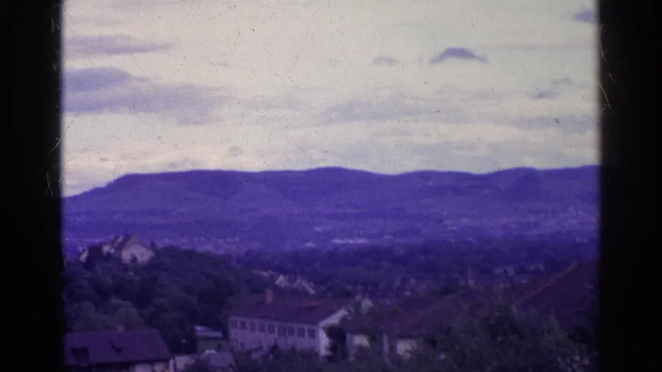 1961 home-movie view across a Swiss valley, with houses and trees below a cloudy sky and purple-blue mountain ridge.